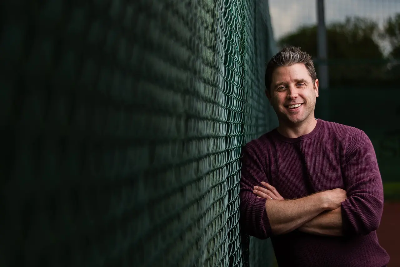 Smilende mand lænet op ad kædehegn på tennisbane