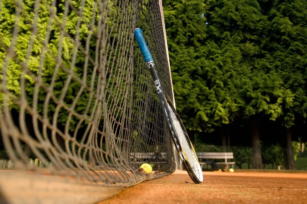 Tennisracket lænet mod nettet og en tennisbold på grusbane